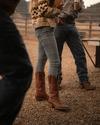 People standing on gravel outdoors, wearing jeans and cowboy boots. One person holds a can, and fencing and hills are visible in the background.