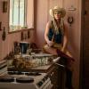 A woman wearing a cowboy hat, denim vest, and boots sits on a kitchen counter holding a bottle, with canning jars and rustic decor around her.