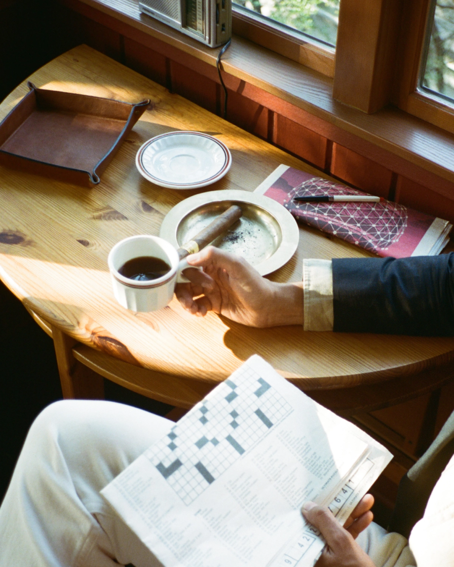 A person holds a cup of coffee and a crossword puzzle at a wooden table with an ashtray and a cigar.