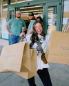 A woman smiles and holds multiple large shopping bags labeled "Tecovas" outside a store. Two men stand in the background.