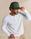 Man in a white Henley shirt and beige pants adjusts a green cap while seated against a neutral background.