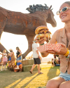 Woman enjoying a large burger at an outdoor festival, with a giant horse sculpture and crowd in the background.