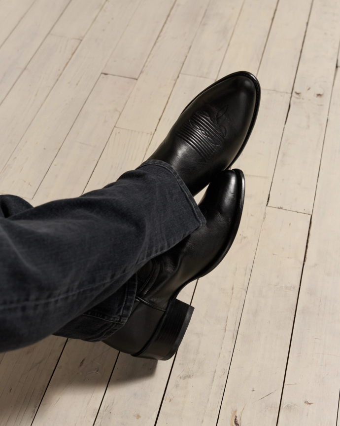 Person wearing black leather boots and dark jeans, seated on light wooden floor.