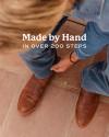 Person in blue jeans and brown boots adjusting their pant leg, with text overlay stating "Made by Hand in Over 200 Steps.