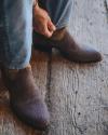 Close-up of a person's lower legs and feet, wearing jeans and textured brown cowboy boots on a wooden floor.