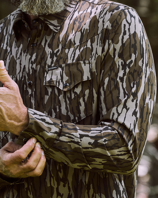 Person adjusts the cuff of a long-sleeved camouflage shirt with a chest pocket, outdoors. Only the torso and hands are visible.