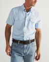 A man wearing a light blue short-sleeve button-up shirt with front pockets, blue jeans, and a black belt, standing against a plain background.