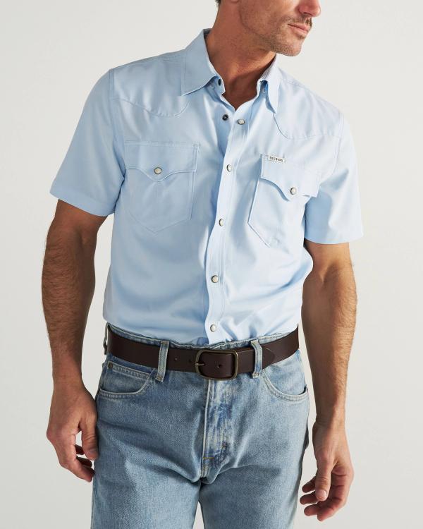 A man wearing a light blue short-sleeve button-up shirt with front pockets, blue jeans, and a black belt, standing against a plain background.