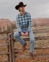 A man in a plaid shirt, jeans, cowboy hat, and boots sits on a wooden fence in a rural landscape with red rock cliffs in the background.