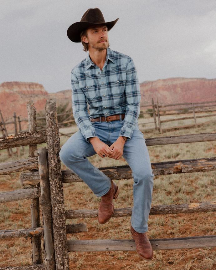 A man in a plaid shirt, jeans, cowboy hat, and boots sits on a wooden fence in a rural landscape with red rock cliffs in the background.