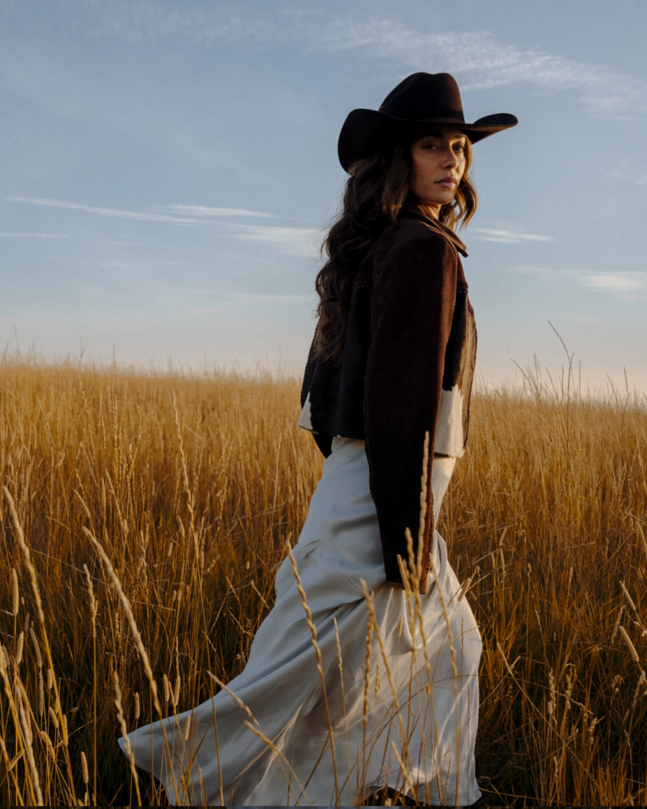 A woman wearing a cowboy hat and dark jacket stands in a tall grass field at sunset.