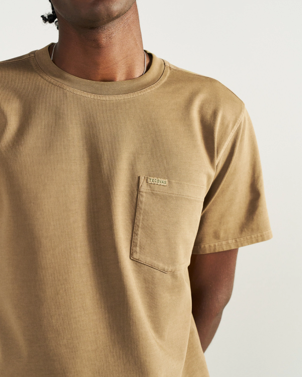 A person wearing a tan short-sleeve T-shirt with a chest pocket and small label, photographed from the shoulders to the waist against a plain background.
