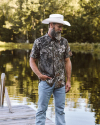 A man in a cowboy hat, camo shirt, and jeans stands on a wooden dock by a lake, with trees in the background.