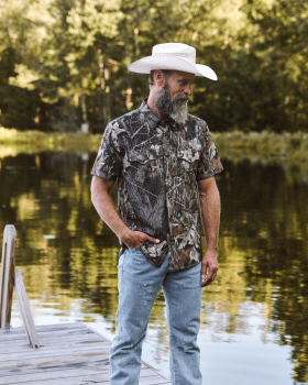 A man in a cowboy hat, camo shirt, and jeans stands on a wooden dock by a lake, with trees in the background.