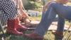 A group of people in jeans and boots is sitting on grass and chairs by a riverside. One person has a bottle in their hand, and a cooler is visible in the background.
