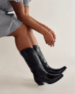 Person wearing a black-and-white checkered dress and black cowboy boots, sitting with arms resting on knees and fingers adorned with a ring and bracelet.