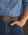 A person wearing a blue denim shirt and jeans with a brown belt featuring an ornate buckle engraved with an eagle.
