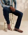 A person wearing a striped shirt, dark jeans, and brown cowboy boots sits on a wooden stool.