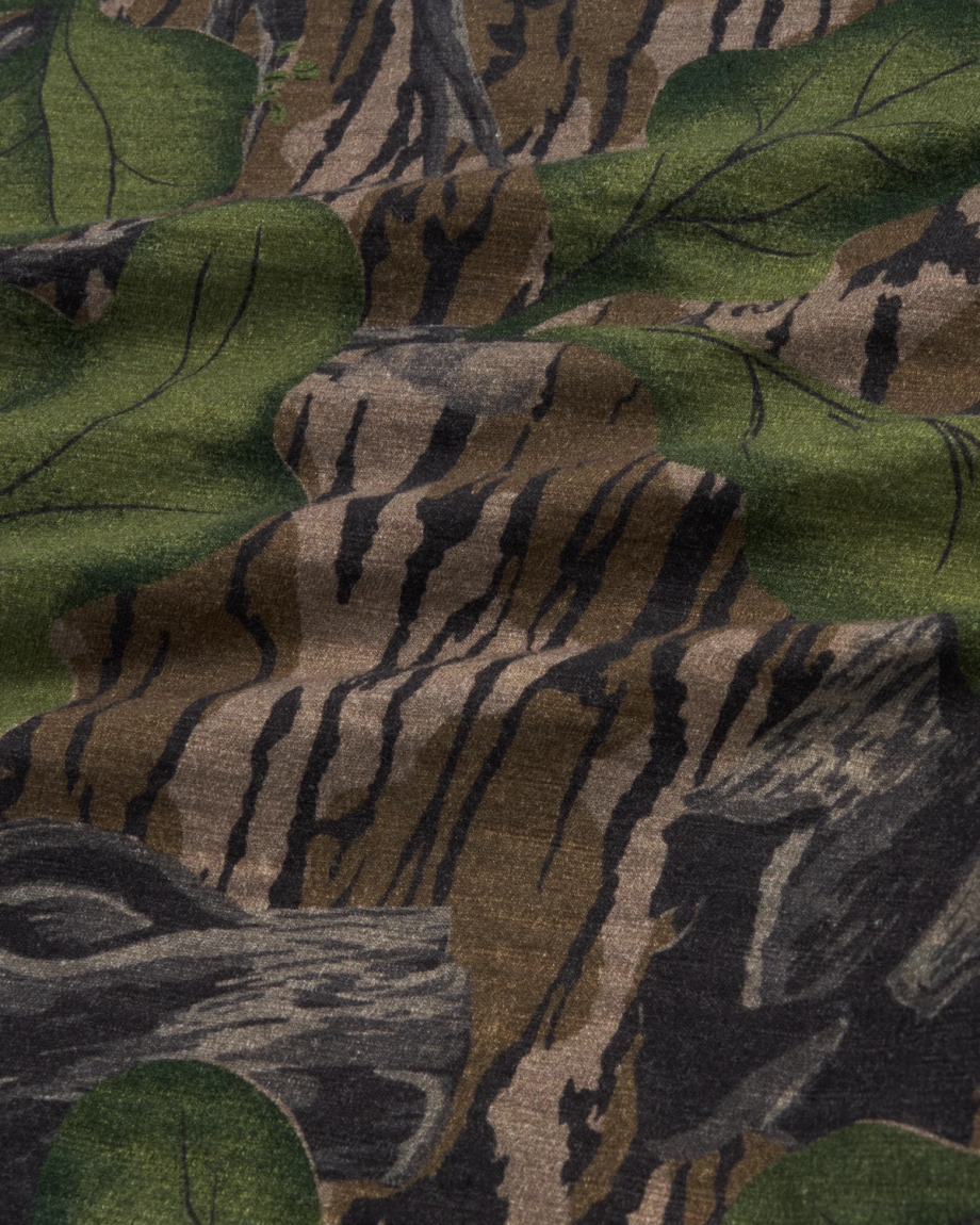 A close-up view of camouflage fabric with a pattern resembling tree bark, branches, and green leaves.