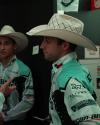 Two men in cowboy hats and teal-patterned shirts stand indoors, one facing forward and the other in profile, with signs and posters visible in the background.