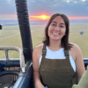 A person smiles in a hot air balloon basket at sunrise, with fields and another balloon visible in the background.