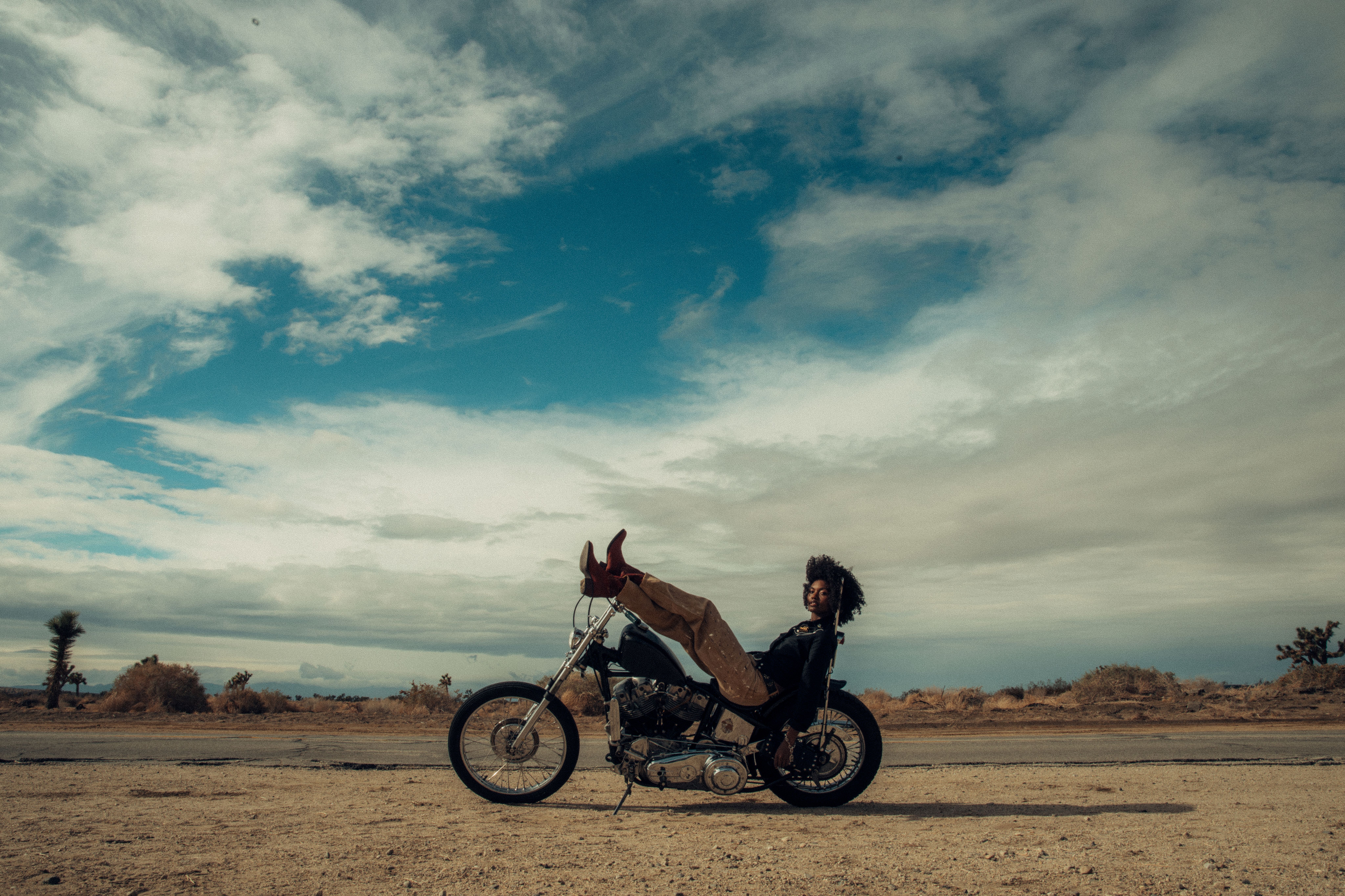 A person reclines on a motorcycle in a desert landscape, with legs propped up on the handlebars and a cloudy sky overhead.