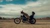 A person reclines on a motorcycle in a desert landscape, with legs propped up on the handlebars and a cloudy sky overhead.