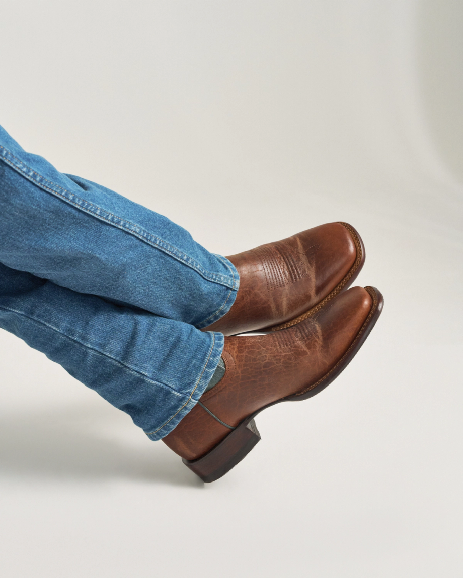 The Doc in Burnt Umber / Blue Cowhide. A person wearing blue jeans and The Doc Cowhide brown leather cowboy boots with legs crossed against a plain background.