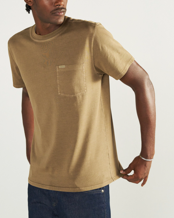 A person wearing a tan short-sleeve t-shirt with a chest pocket and dark blue jeans stands against a plain background.