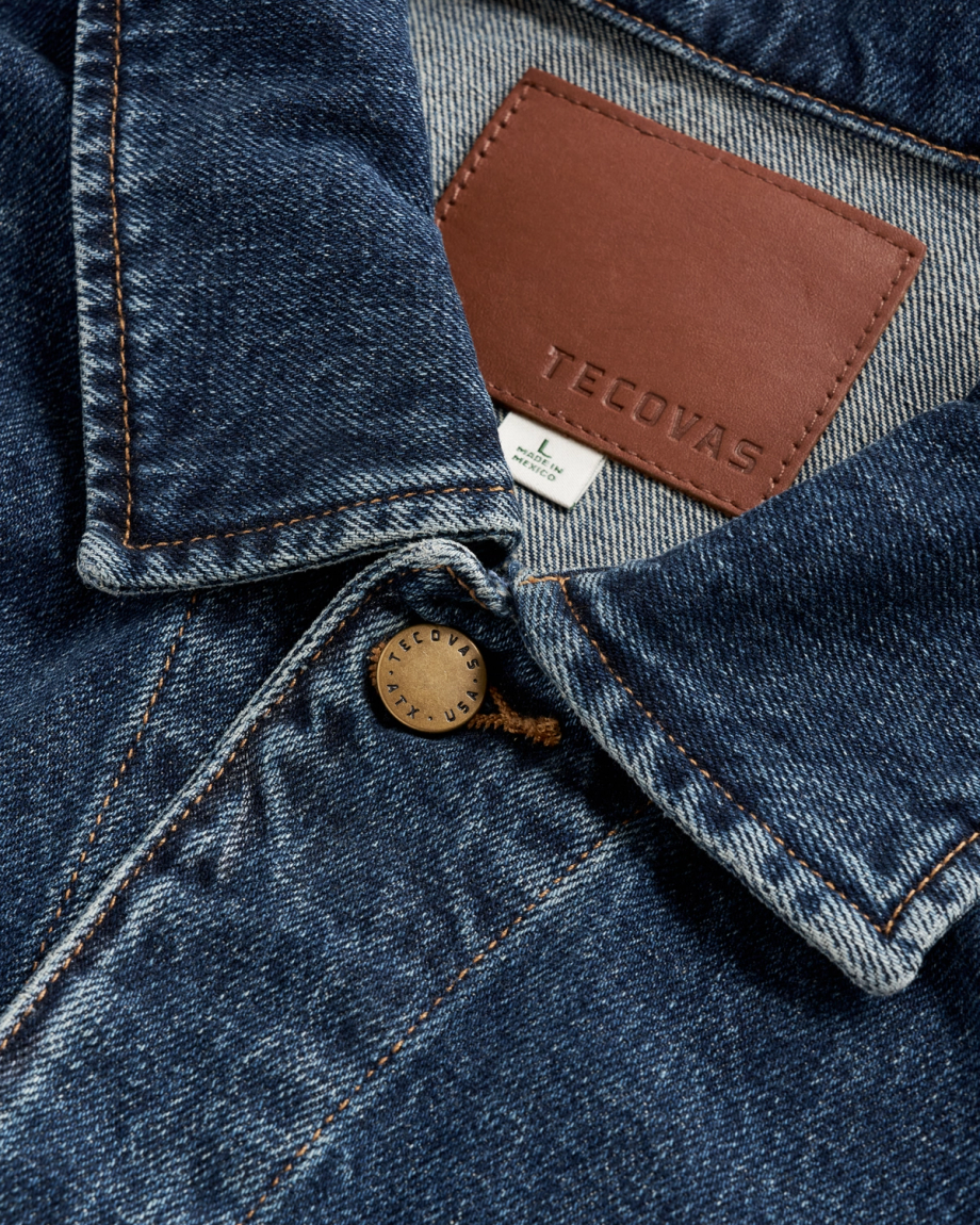 Close-up of a blue denim jacket collar featuring a brown leather Tecovas brand patch, a size Large tag made in Mexico, and a brass button stamped with “Tecovas ATX USA.”.