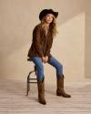 Woman sitting on a metal stool with cowboy hat, fringe blazer, denim jeans and brown cowgirl boots.