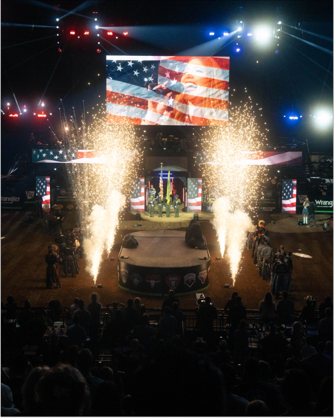 Two cowboys on a stage with fireworks and a large screen above displaying their image at an indoor rodeo event. Bright lights and audience in the background.