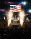 Two cowboys on a stage with fireworks and a large screen above displaying their image at an indoor rodeo event. Bright lights and audience in the background.