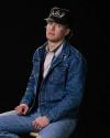 A person wearing a denim jacket, jeans, and a black cap with an antler design sits on a stool against a black background.