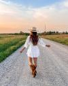 A woman walking down a dirt road in a white dress and cowboy boots.