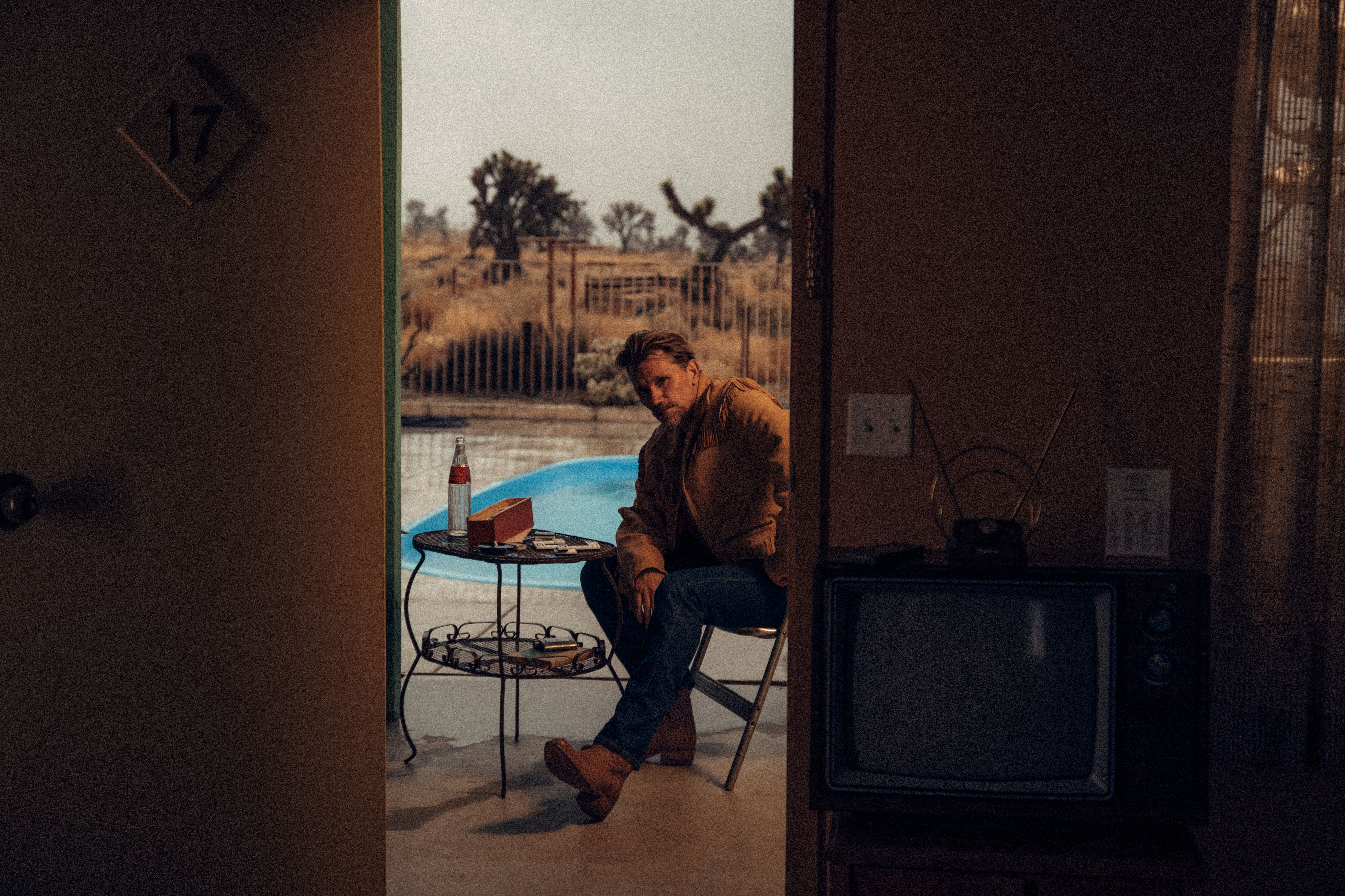 A man sits at a table by a pool outside a motel room, with a bottle, a takeout box, and an old television visible in the foreground.