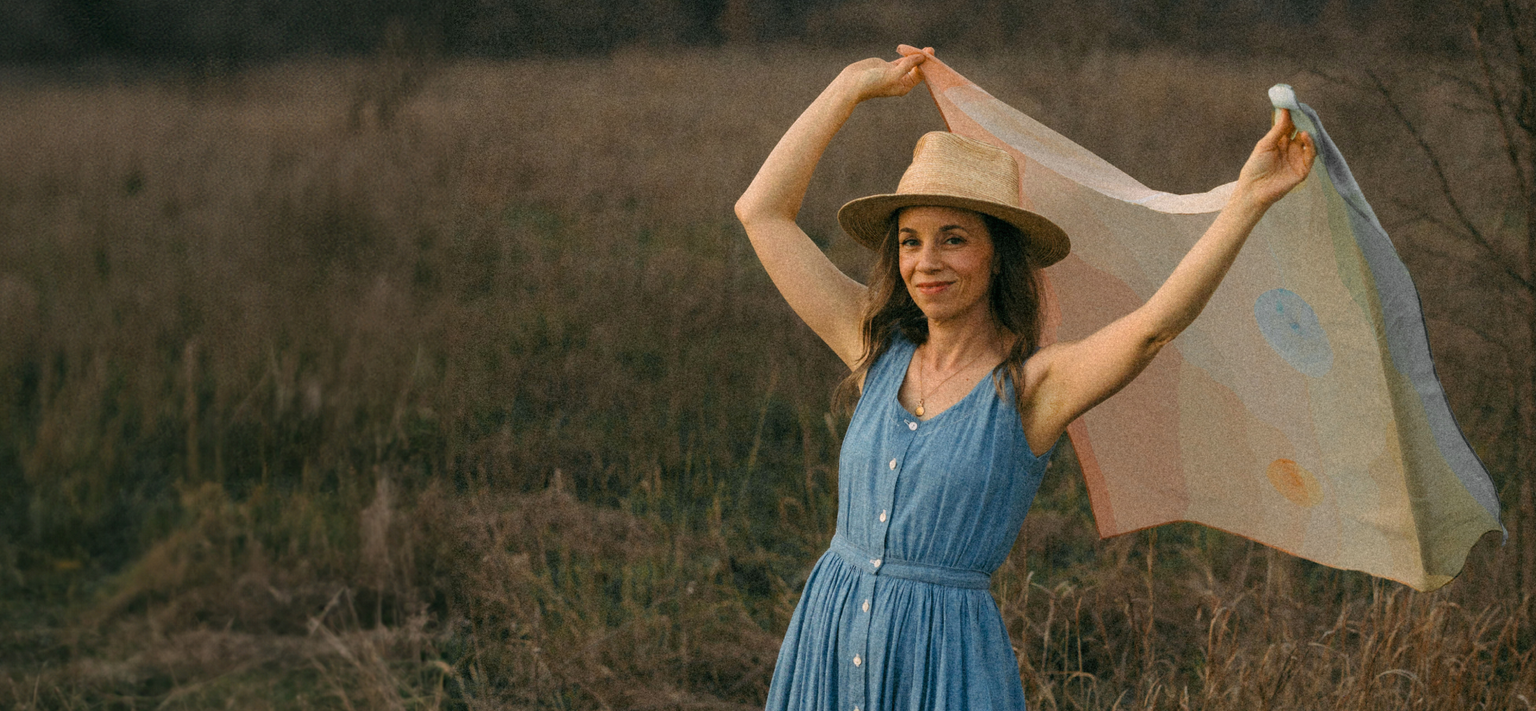 A woman in a straw hat and blue dress smiling, holding up a light scarf in a grassy field at dusk.