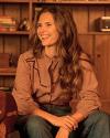 A woman with long wavy hair sits and smiles, wearing a checked shirt and jeans in a cozy room with wooden shelves and books in the background.