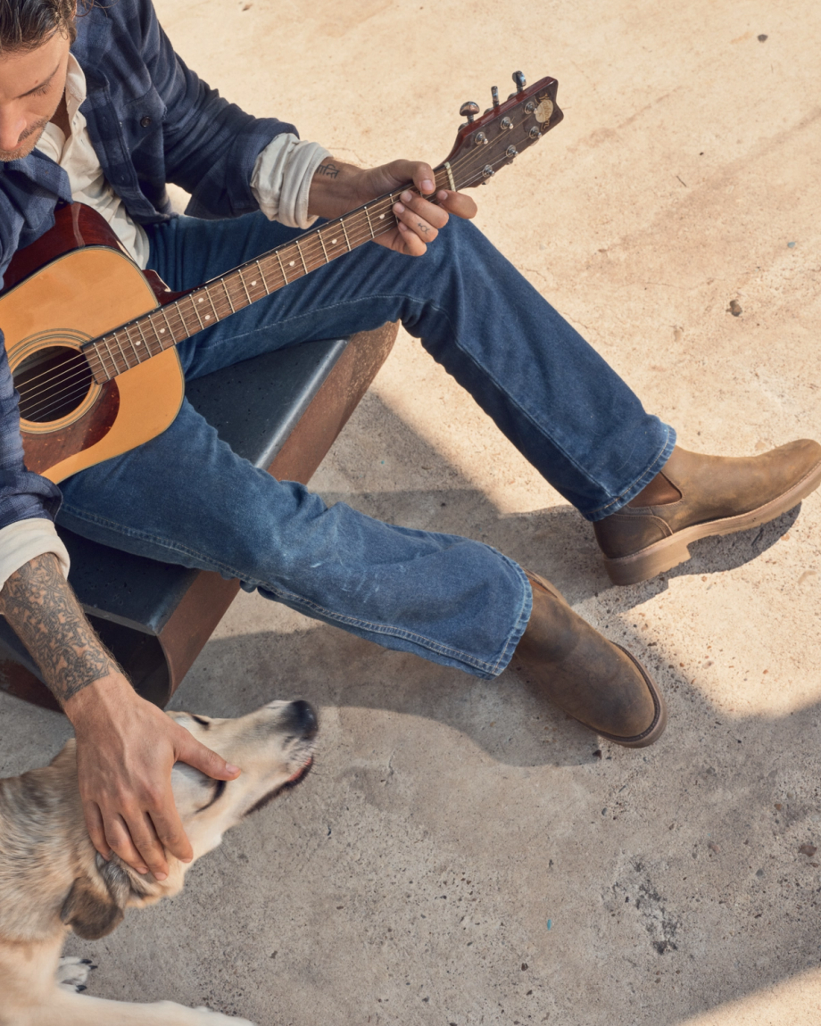 A man sits on a bench playing guitar with his loyal dog beside him, his The Rugged Chelsea Cowhide canteen resting at his feet.
