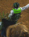 A rodeo rider wearing a helmet and protective vest rides a bull in an arena, with one arm raised and green fringe on his gear.