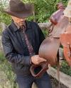 A person wearing a dark hat and jacket is inspecting a brown leather saddle hanging on a wooden fence.