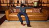 A person in a cowboy hat, denim shirt, blue jeans, and boots sits relaxed on a brown leather sofa in front of a display of boots.