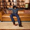 A person in a cowboy hat, denim shirt, blue jeans, and boots sits relaxed on a brown leather sofa in front of a display of boots.