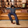 A person in a cowboy hat, denim shirt, blue jeans, and boots sits relaxed on a brown leather sofa in front of a display of boots.