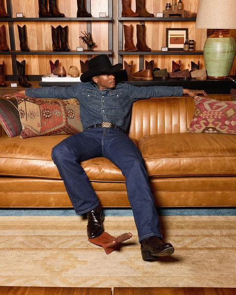 A person in a cowboy hat, denim shirt, blue jeans, and boots sits relaxed on a brown leather sofa in front of a display of boots.