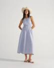 The Carmen Paloma in Pearl Ostrich. Woman wearing a sleeveless blue and white gingham dress, white sandals, a wide-brimmed hat, and a delicate pearl necklace, standing with hands behind her back against a plain background.
