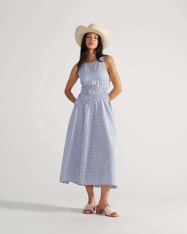 The Carmen Paloma in Pearl Ostrich. Woman wearing a sleeveless blue and white gingham dress, white sandals, a wide-brimmed hat, and a delicate pearl necklace, standing with hands behind her back against a plain background.