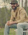 Man wearing a beige button-up shirt, blue jeans, and a green cap sitting outdoors and smiling. Trees blurred in the background.