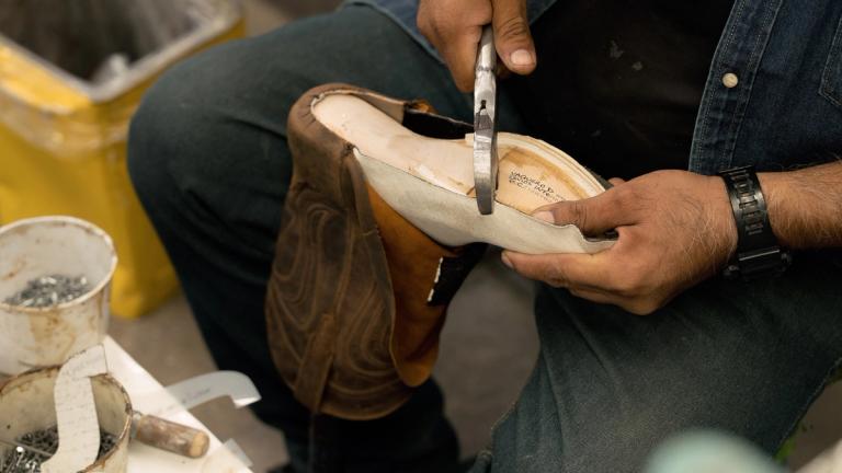 A person uses pliers to work on a leather boot, holding the sole in place. Tools and small containers are visible nearby.