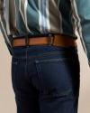 A person wearing dark blue jeans with a brown leather belt and a striped button-down shirt, shown from the waist down, backside view.